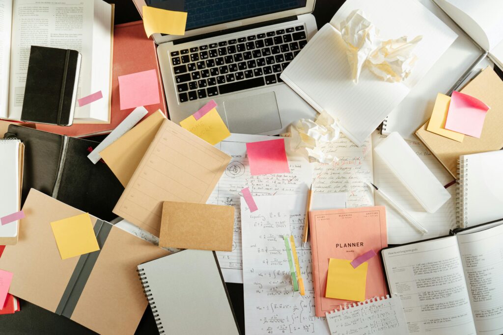 Flatlay of a messy study desk with laptop, notebooks, and sticky notes, showcasing the creative process.